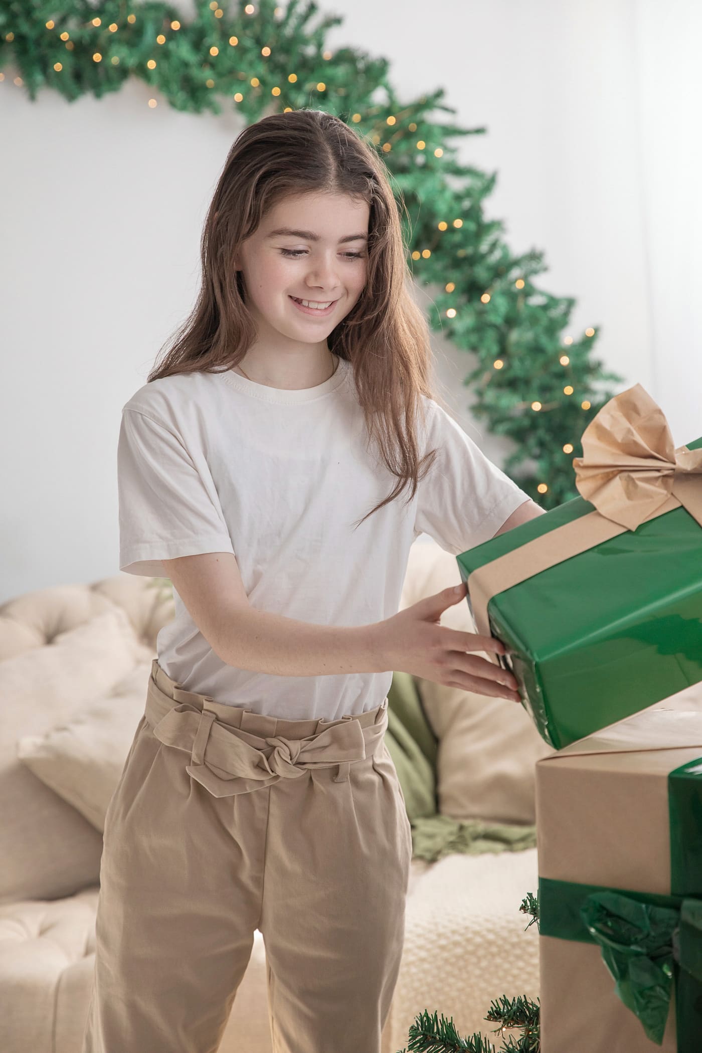 Older girl in a white t-shirt and tan trousers holding a green gift in the Christmas studio