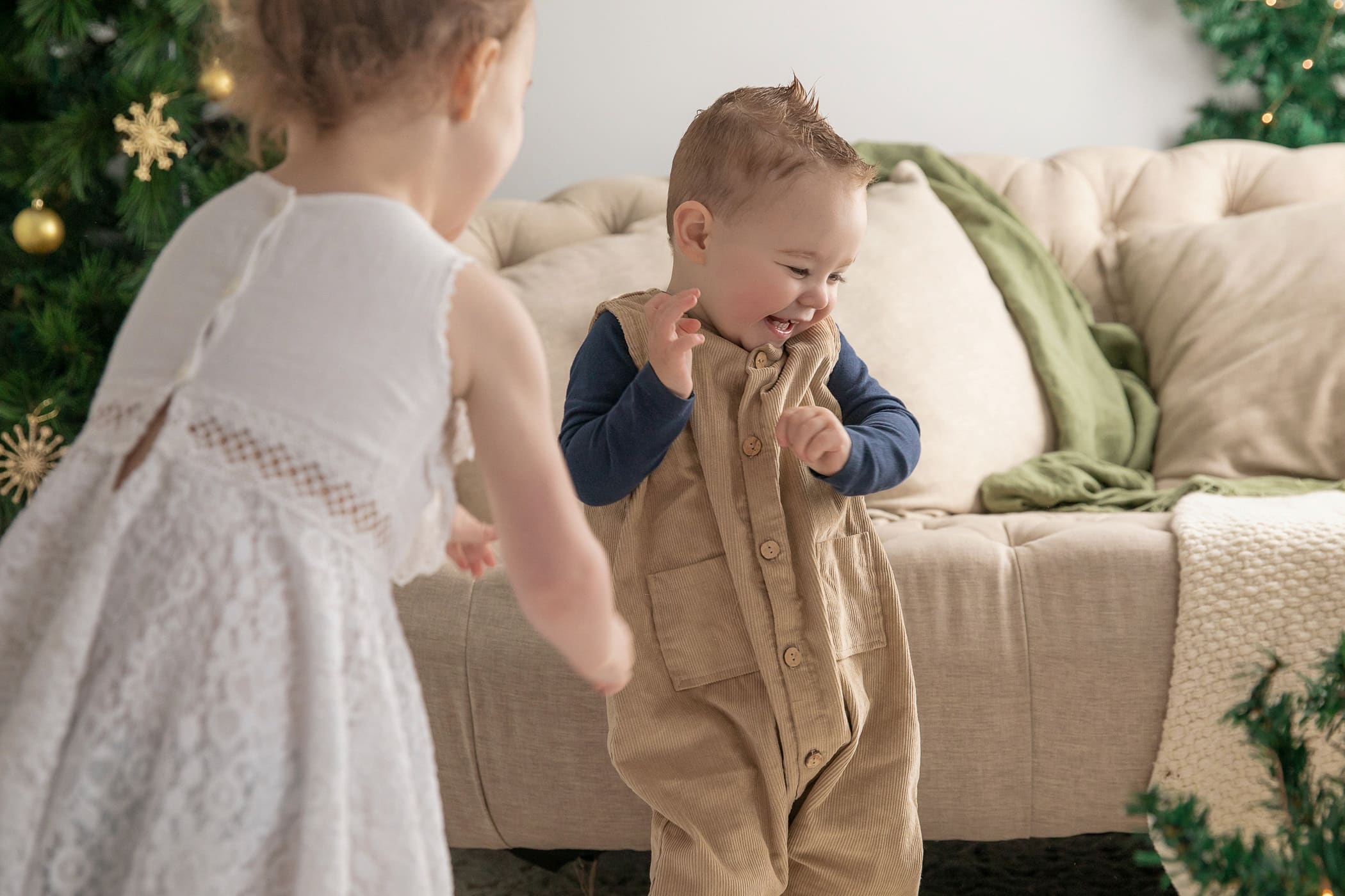 Toddler in tan overalls laughing beside a girl in a white lace dress in the Christmas studio