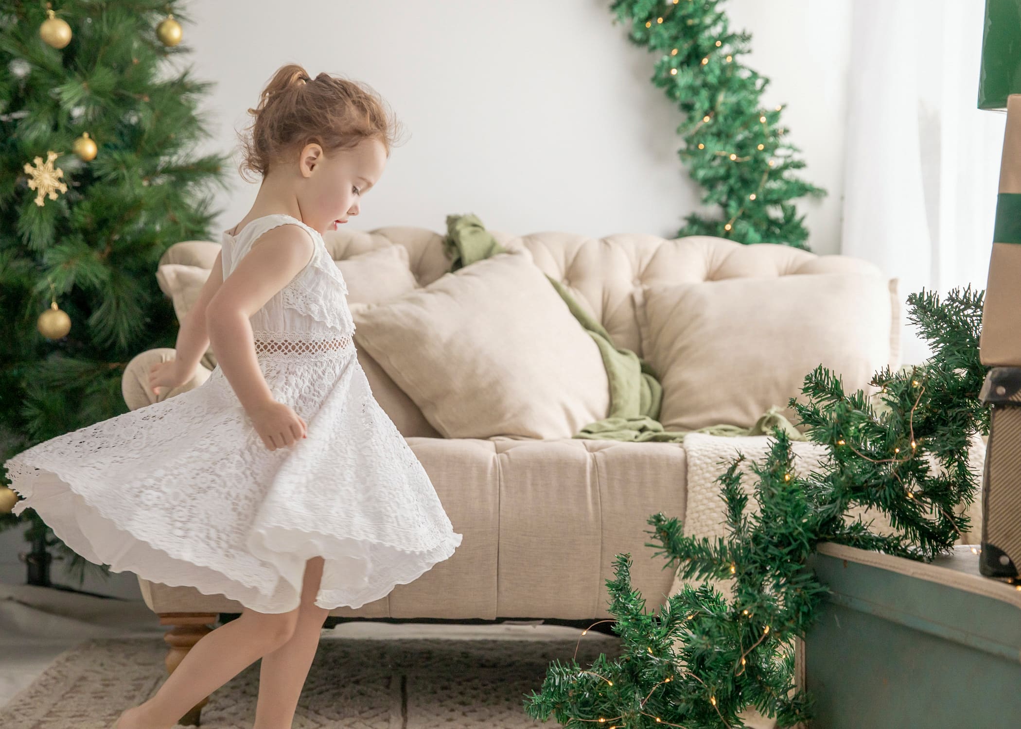 Young girl in a white lace dress twirling beside a Christmas sofa and tree
