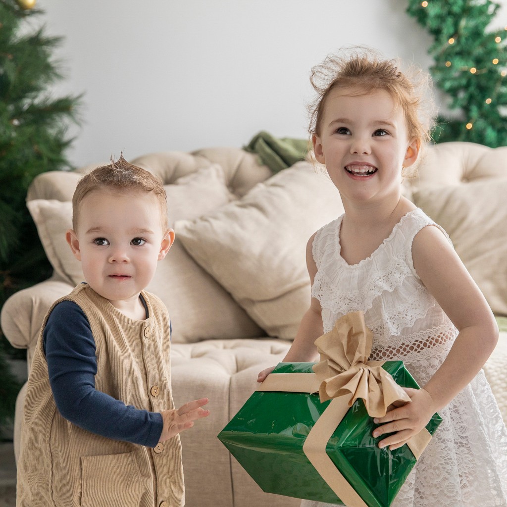Young boy in corduroy overalls and a girl in a white lace dress holding a green gift in the Christmas studio