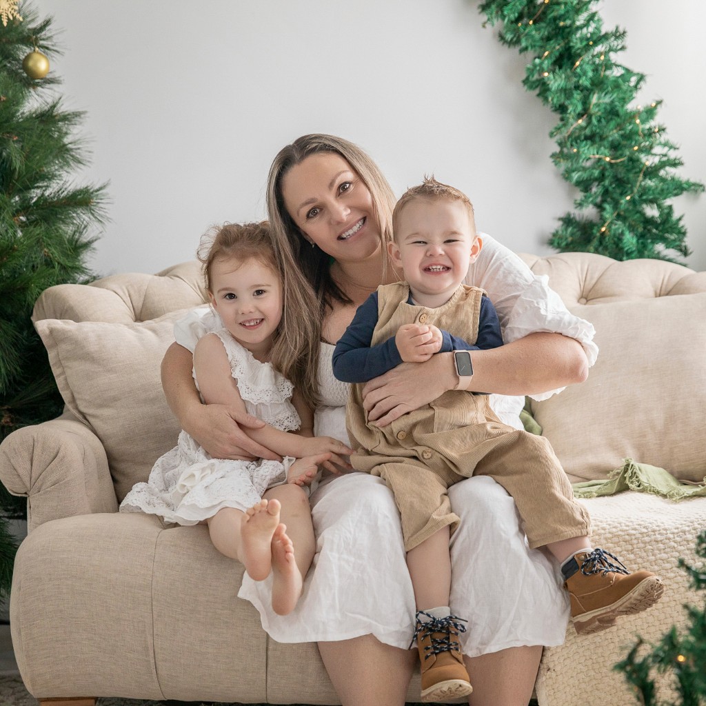 Mother and two young children seated on a tufted sofa between a Christmas tree and a lit pine garland