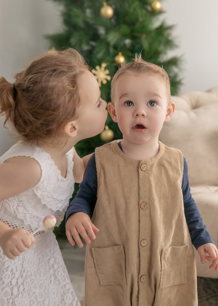 Young girl in a white lace dress kissing a toddler in corduroy overalls in front of a Christmas tree