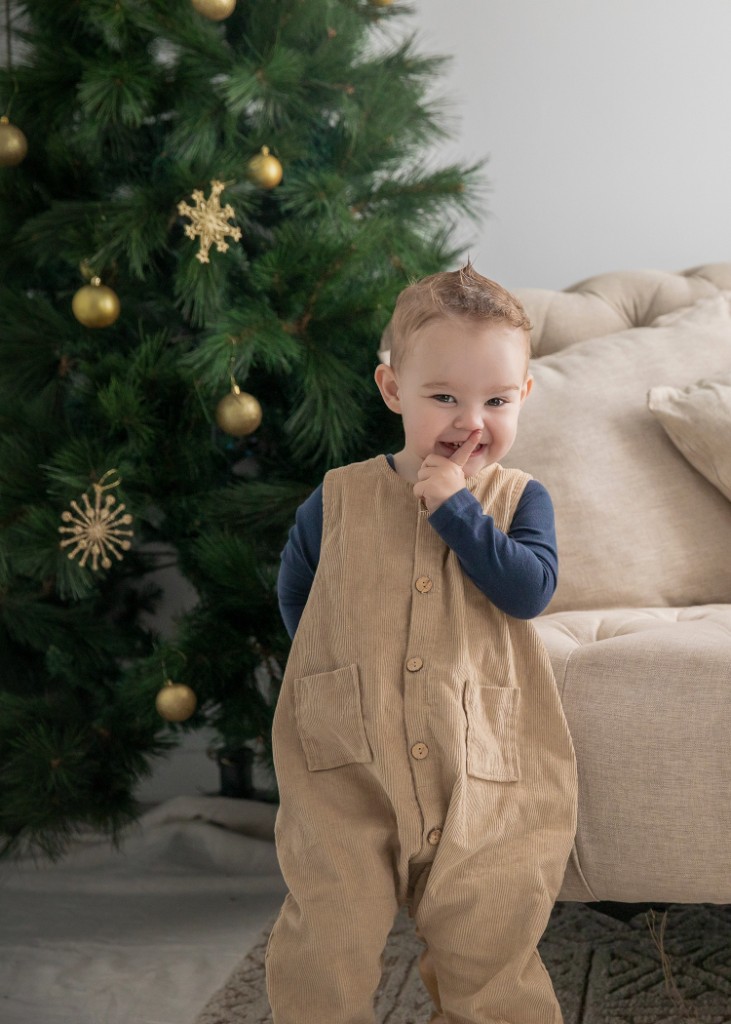 Young child in tan corduroy overalls standing beside a Christmas tree in the studio