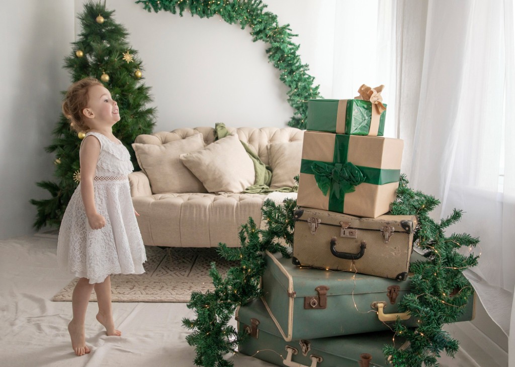 Young girl in a white lace dress looking up beside a Christmas sofa, gift stack and vintage suitcases