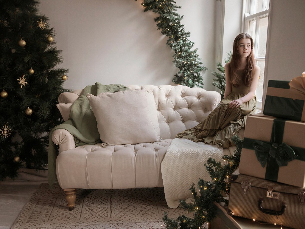 Young girl in an olive dress sitting on a tufted sofa by a window in a Christmas studio set