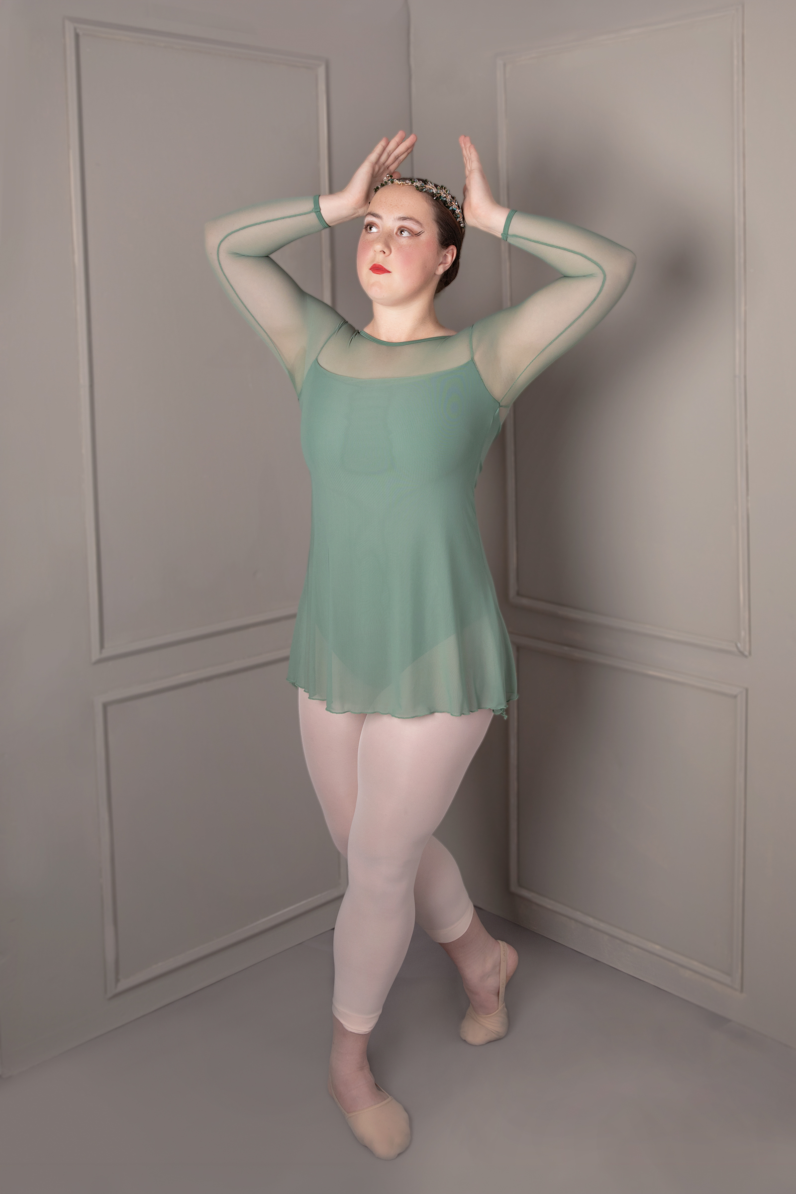 Young dancer in a sage-green costume and jeweled headpiece posing in a studio corner