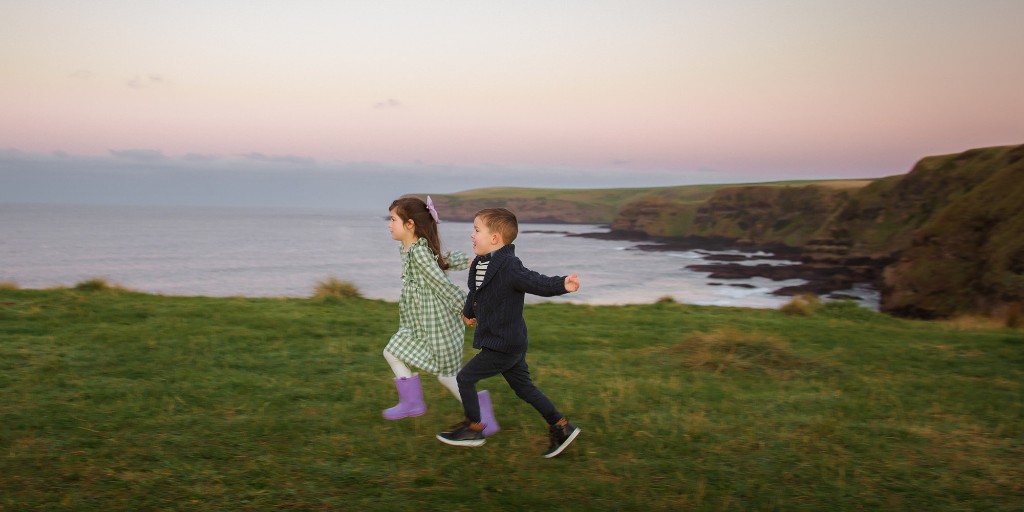 Two children running across a grassy clifftop above the ocean at dusk