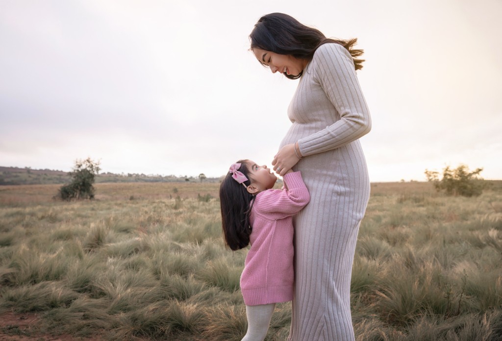Expectant mother in a field with a young child reaching toward her bump