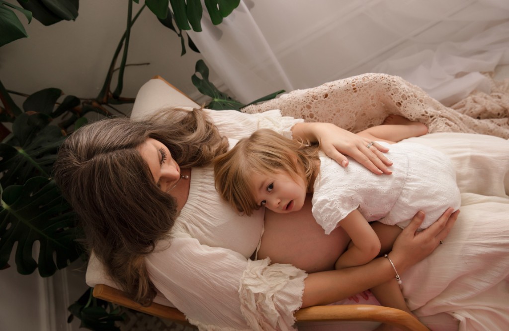 Expectant mother seated with a young child resting against her bump in a bright studio