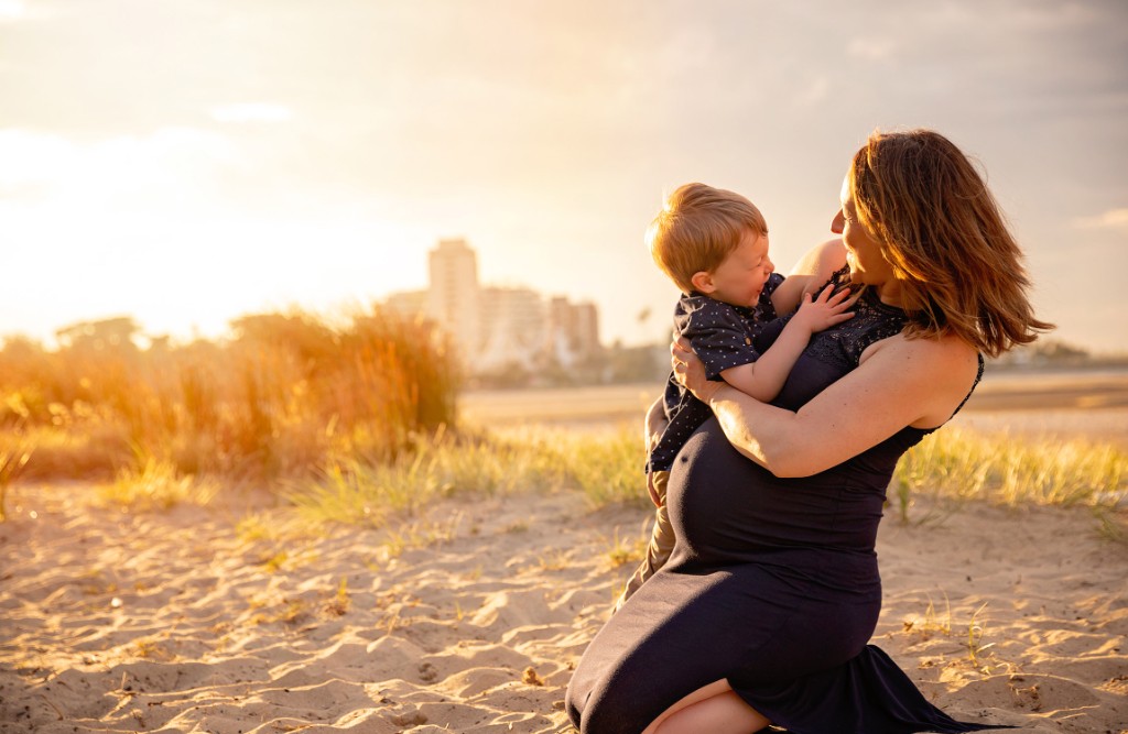 Expectant mother in a navy dress kneeling on a beach at sunset, holding a young child