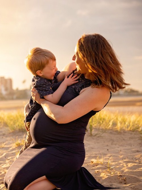 Expectant mother in a navy dress kneeling on a beach at sunset, holding a young child