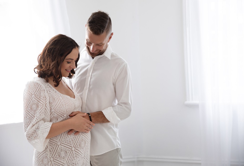 Expectant mother and partner in a bright studio maternity portrait, looking down at her bump