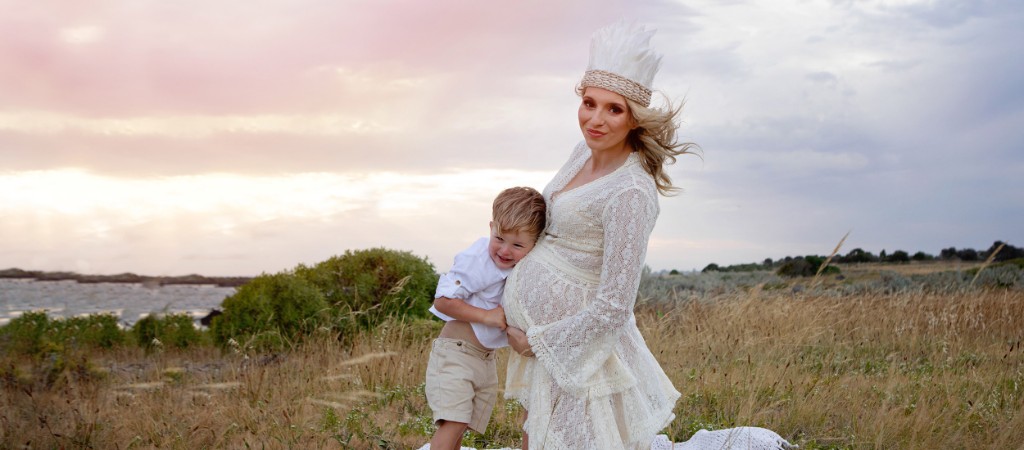 Expectant mother in a white dress with a young child leaning against her bump in a coastal field at sunset